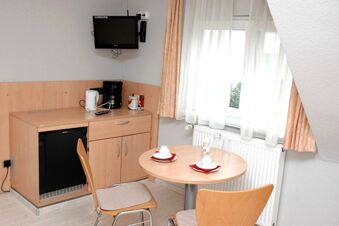 Gem&uuml;tlicher Raum im G&auml;stehaus Boardinghouse Stapf mit kleinem Tisch, zwei St&uuml;hlen, Minik&uuml;hlschrank, TV und Kaffeemaschine am Fenster mit Vorh&auml;ngen.