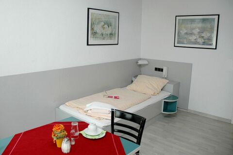 Ein schlicht eingerichtetes Zimmer im G&auml;stehaus Boardinghouse Stapf mit Einzelbett, Tisch mit rotem Tischtuch und zwei gerahmten Bildern an der Wand.