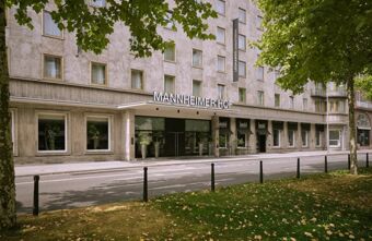 Die Fassade des Leonardo Royal Hotel Mannheim zeigt den modernen Eingangsbereich mit Beschilderung und Fenstern entlang der Stra&szlig;e und gr&uuml;ner Umgebung.