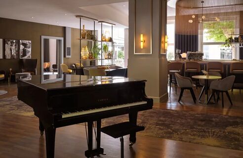 Eleganter Salon mit schwarzem Fl&uuml;gelklavier, moderner Bar und bequemen Sitzgelegenheiten im Leonardo Royal Hotel Mannheim mit warmem Ambiente.