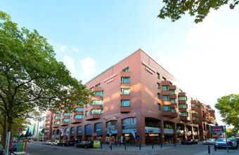 Fassade des mehrst&ouml;ckigen Leonardo Hotel Mannheim City Centre mit roten Backsteinw&auml;nden an einer belebten Stra&szlig;enecke und B&auml;umen im Vordergrund.