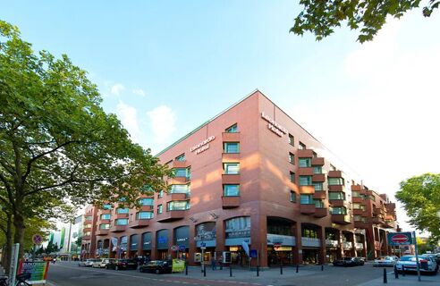 Fassade des mehrst&ouml;ckigen Leonardo Hotel Mannheim City Centre mit roten Backsteinw&auml;nden an einer belebten Stra&szlig;enecke und B&auml;umen im Vordergrund.