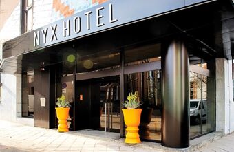 Die Fassade des NYX Hotel Mannheim zeigt einen modernen Eingang mit gro&szlig;en Glasfl&auml;chen und zwei orangenen Pflanzgef&auml;&szlig;en an der Au&szlig;enseite.