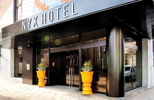 Die Fassade des NYX Hotel Mannheim zeigt einen modernen Eingang mit gro&szlig;en Glasfl&auml;chen und zwei orangenen Pflanzgef&auml;&szlig;en an der Au&szlig;enseite.
