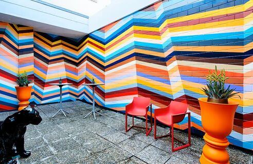 Bunte, geometrisch gemusterte Wandgestaltung mit roten St&uuml;hlen und orangefarbenen Pflanzgef&auml;&szlig;en im Au&szlig;enbereich des NYX Hotel Mannheim.