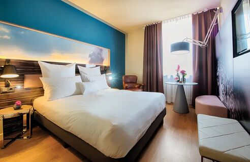 Modern eingerichtetes Hotelzimmer mit gro&szlig;em Bett, Sitzbereich und gro&szlig;em Fenster im NYX Hotel Mannheim f&uuml;r erholsamen Aufenthalt.