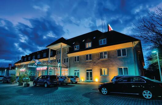 Das beleuchtete Geb&auml;ude des Leonardo Hotel Mannheim-Ladenburg bei Abendd&auml;mmerung mit mehreren geparkten Autos vor dem Eingang.