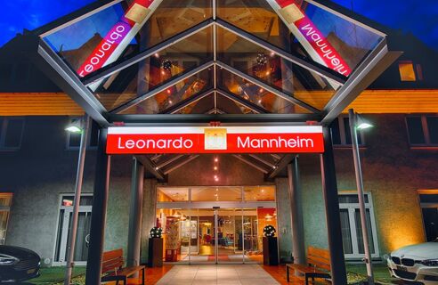 Nachtaufnahme des Eingangsbereichs vom Leonardo Hotel Mannheim-Ladenburg mit beleuchtetem Schild und Glasdach am Hoteleingang.
