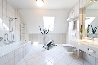 Bright bathroom at Leonardo Hotel Heidelberg-Walldorf with white tiles, bathtub, elliptical trainer, toilet, and sink with mirror and flowers