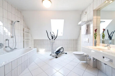 Bright bathroom at Leonardo Hotel Heidelberg-Walldorf with white tiles, bathtub, elliptical trainer, toilet, and sink with mirror and flowers