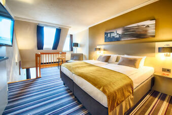 A modern hotel room at Leonardo Hotel Heidelberg-Walldorf with a large bed, striped carpet, armchair, table, and wall artwork