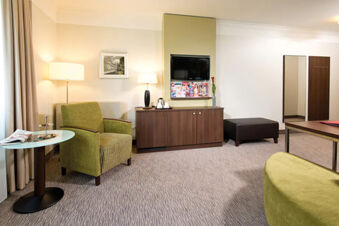 Hotel room at Leonardo Hotel Heidelberg-Walldorf with green armchair, floor lamps, TV mounted on wall, and wooden cabinet
