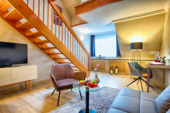 Modern living area with wooden staircase, brown armchair, glass coffee table, TV, and workspace in Leonardo Hotel Heidelberg-Walldorf