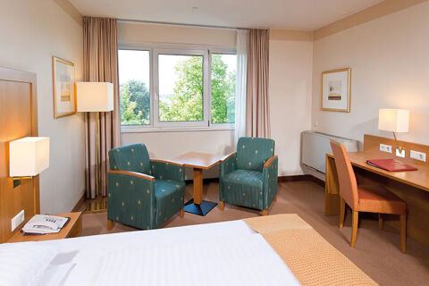 A hotel room at Leonardo Hotel Heidelberg-Walldorf with two green armchairs, a small wooden table, a bed, and a desk with a chair