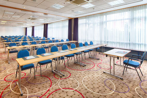 Conference room at Leonardo Hotel Heidelberg-Walldorf with rows of tables and blue chairs facing windows with sheer curtains
