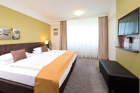 Hotel room at Leonardo Hotel Heidelberg-Walldorf with two single beds, mustard wall, TV, bench, and large window with curtains