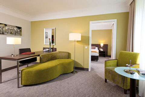 A modern hotel room with olive green sofa, armchair, glass table, desk with chair and a floor lamp at Leonardo Hotel Heidelberg-Walldorf