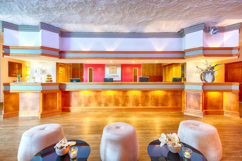 Reception area of Leonardo Hotel Heidelberg-Walldorf with wood paneling, three computer monitors, and three beige stools by round tables