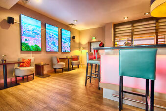 Lobby seating area with beige chairs, red pillows, illuminated bar stools, and three large cityscape screens at Leonardo Hotel Heidelberg-Walldorf