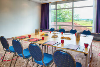 Meeting room at Leonardo Hotel Heidelberg-Walldorf with blue chairs, wooden tables, red mugs, and a large window showing trees outside