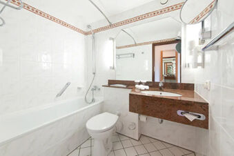 Bathroom in Leonardo Hotel Heidelberg-Walldorf with white tiled walls, bathtub, toilet, brown granite sink counter, and large mirror
