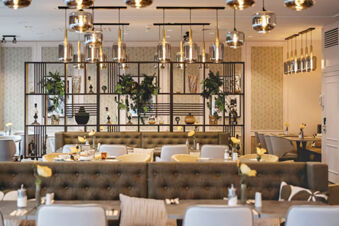 Dining area with tufted brown benches, white and yellow chairs, decorative shelves, and hanging lights at Leonardo Hotel Heidelberg City Center
