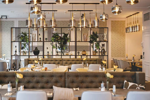 Dining area with tufted brown benches, white and yellow chairs, decorative shelves, and hanging lights at Leonardo Hotel Heidelberg City Center