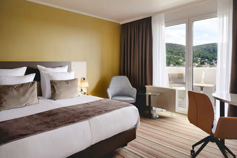 Hotel room at Leonardo Hotel Heidelberg City Center with a made bed, armchair, desk chair, and balcony overlooking hills and houses