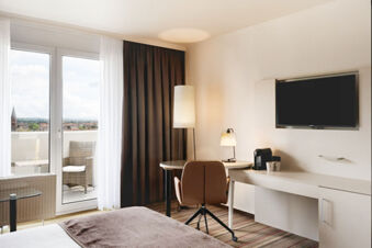 Hotel room at Leonardo Hotel Heidelberg City Center with a bed, desk, chair, TV, floor lamp, and balcony with outdoor furniture visible
