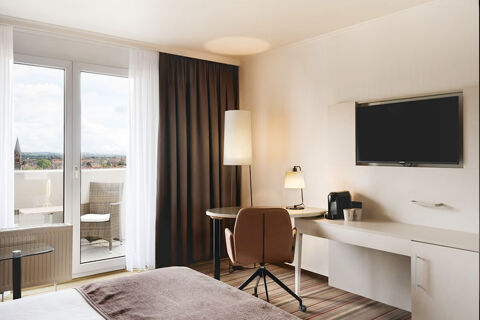Hotel room at Leonardo Hotel Heidelberg City Center with a bed, desk, chair, TV, floor lamp, and balcony with outdoor furniture visible