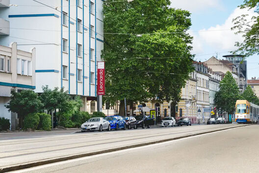Street view of Leonardo Hotel Heidelberg City Center with parked cars, green trees, and a tram on tram tracks under a partly cloudy sky