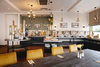 Buffet breakfast area with food warmers, juice dispensers, plates, and yellow chairs in Leonardo Hotel Heidelberg City Center