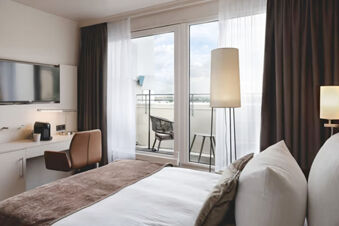 Modern hotel room at Leonardo Hotel Heidelberg City Center with bed, desk, chair, TV, floor lamp, and balcony seating visible through glass doors