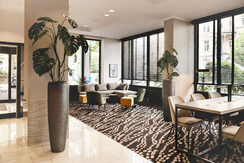 A spacious seating area in Leonardo Hotel Heidelberg City Center with a large plant, modern chairs, a sofa, and windows with blinds
