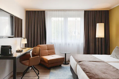 Hotel room at Leonardo Hotel Heidelberg City Center showing a bed with brown runner, leather chair, desk, armchair, and large window with curtains