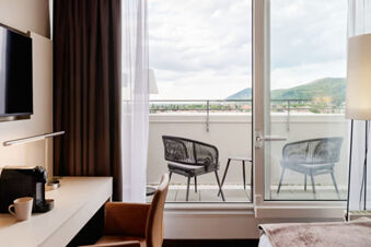 Interior view of a Leonardo Hotel Heidelberg City Center room showing a desk, chair, TV, coffee machine, balcony with two chairs and table, and hills outside