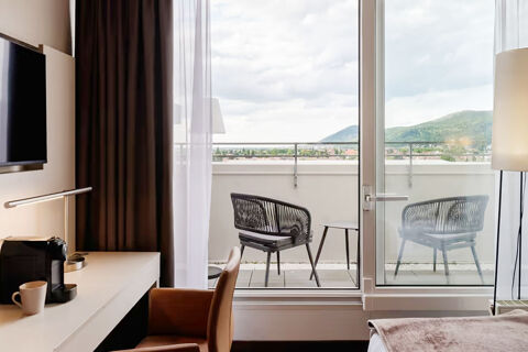 Interior view of a Leonardo Hotel Heidelberg City Center room showing a desk, chair, TV, coffee machine, balcony with two chairs and table, and hills outside