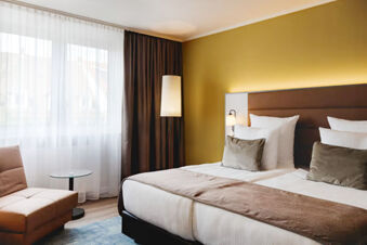 Hotel room at Leonardo Hotel Heidelberg City Center featuring a double bed with pillows, a floor lamp, an armchair, and a window with curtains