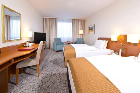 Two beds with white linens and orange covers in a room at Leonardo Hotel Heidelberg with desk, TV, two chairs, and lamps
