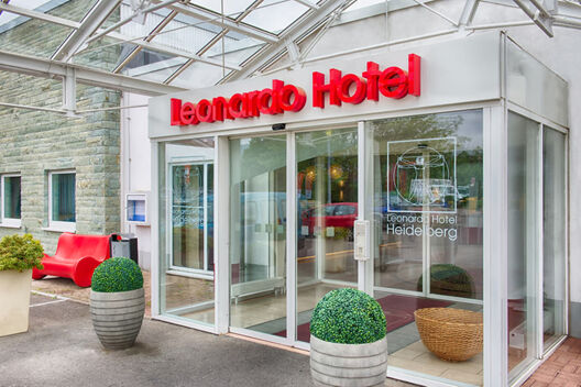 Entrance of Leonardo Hotel Heidelberg with glass doors, red signage, two large potted plants, and a red bench outside the building