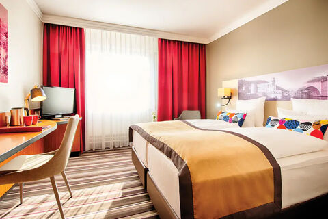 Hotel bedroom at Leonardo Hotel Heidelberg with twin beds, colorful pillows, red curtains, desk, chair, and TV visible