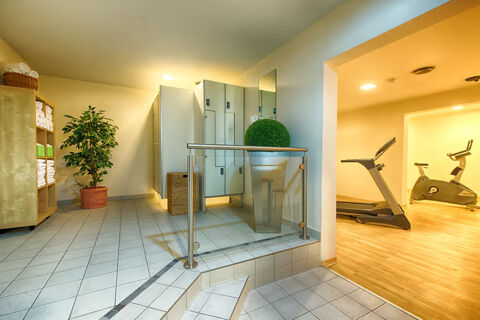 Locker area with tiled floors, potted plants, wooden shelves with towels, and adjacent gym equipment at Leonardo Hotel Heidelberg