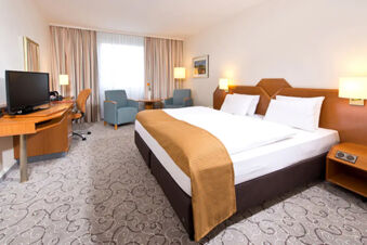 Hotel room at Leonardo Hotel Heidelberg featuring a large bed with a brown throw, two chairs by a window, and a desk with a TV and chair