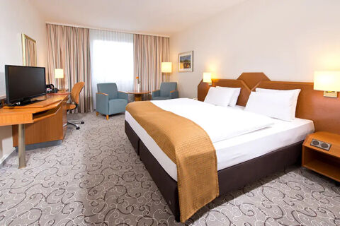 Hotel room at Leonardo Hotel Heidelberg featuring a large bed with a brown throw, two chairs by a window, and a desk with a TV and chair
