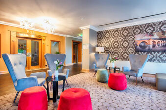 Lounge area in Leonardo Hotel Heidelberg with gray armchairs, red and green ottomans, small black tables, and patterned wallpaper