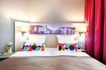 Double bed with colorful pillows, white sheets, wall lamps, red curtains, and a photo headboard at Leonardo Hotel Heidelberg