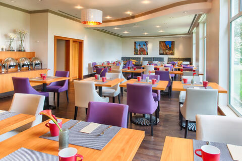 Dining area at Leonardo Hotel Heidelberg with wooden tables, purple and white chairs, red mugs, and large windows on one side