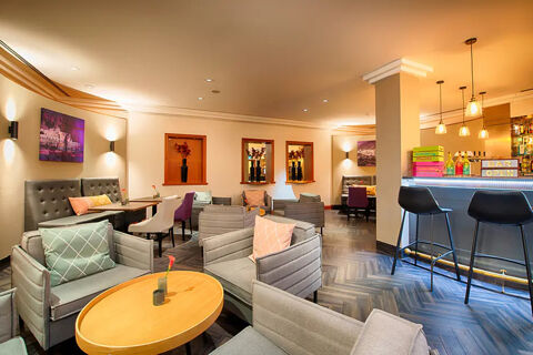 Modern lounge area with gray chairs, colorful cushions, wooden tables, and a bar at Leonardo Hotel Heidelberg