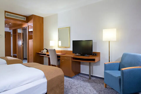 Hotel room at Leonardo Hotel Heidelberg with two single beds, a wooden desk with a TV, chair, blue armchair, and floor lamp