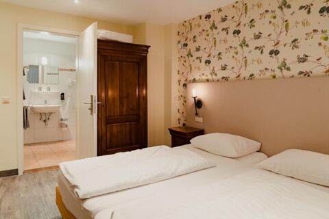 White bedding on twin beds, floral wallpaper, wooden closet, nightstand with lamp, and bathroom visible at Gasthaus Backmulde - Hotel